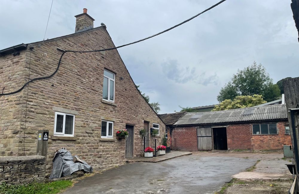 A stone built farm house with a brick outbuilding with a corrugated iron roof on the other side of a yard