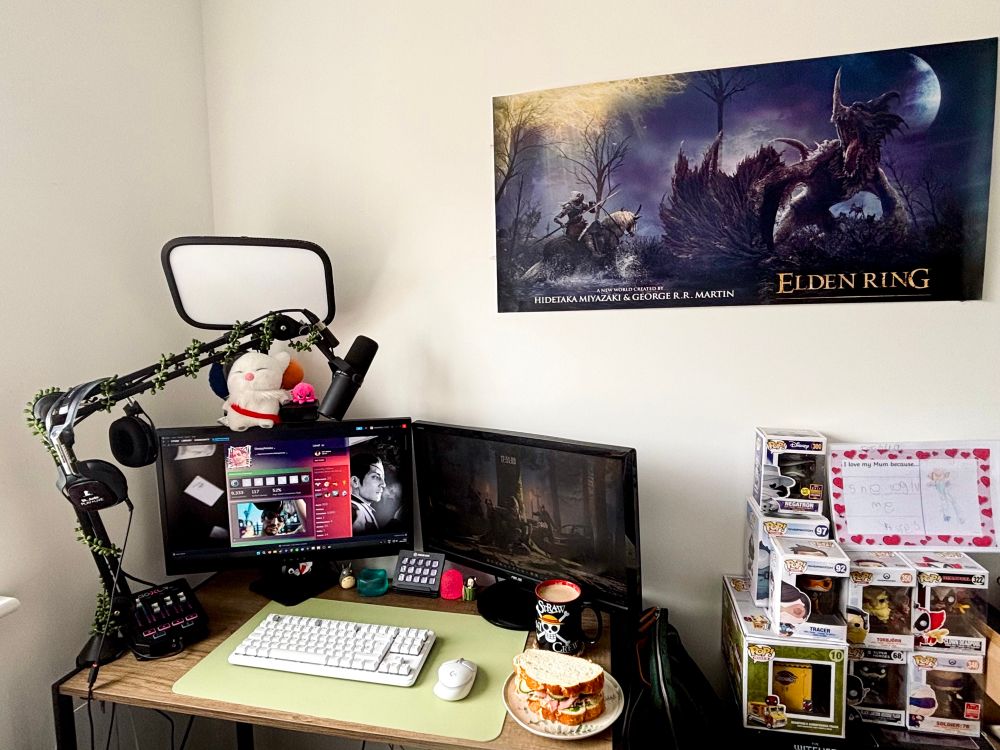 a desk with two monitors side by side with streaming audio systems, key light, keyboard, mouse, sandwich and coffee on it. beside the desk is a stack of funko pops and above is an elden ring poster