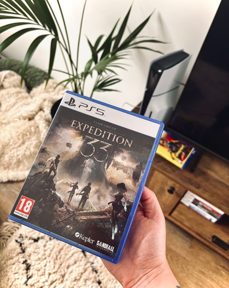 the physical game clair obscur expedition 33 on ps5 held in front of a wooden tv unit with a tv, ps5 and various games on it. bedside it is a kentia palm and white blanket