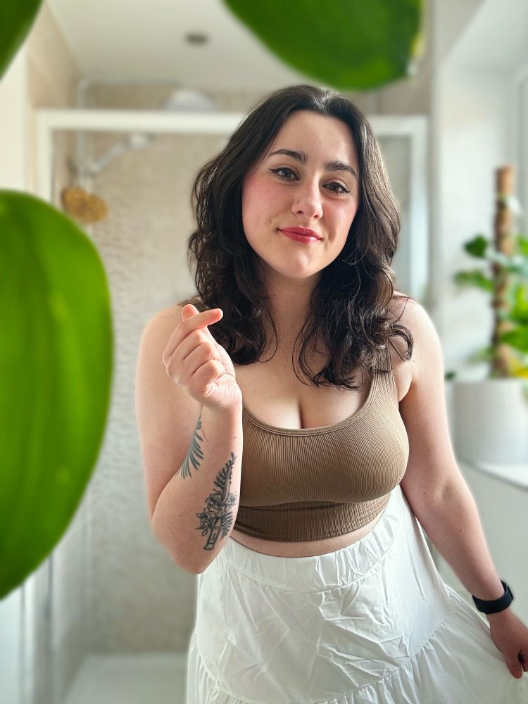 nosku posing in a white tiered skirt, coffee crop vest top in her on suite with her plant framing the selfie 