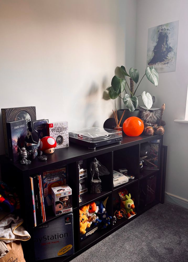 a kallax shelving unit on its side with various items such as old consoles, plushies and statues. on top of it are from soft and hollow knight books, a vinyl player, monstera, an orange orb light and a lord of the rings build a bear. on the wall is a last of us displate of ellie playing a guitar