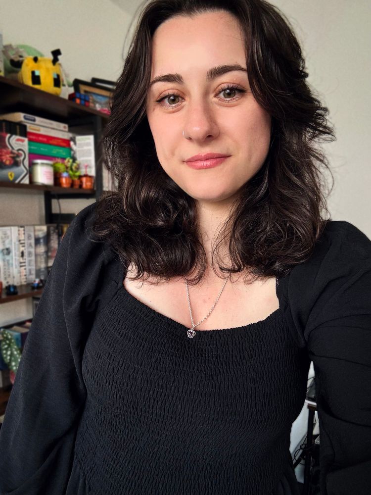 nosku in a black jumpsuit smiling with a silver chain and nose ring. she is stood in front of a book shelf