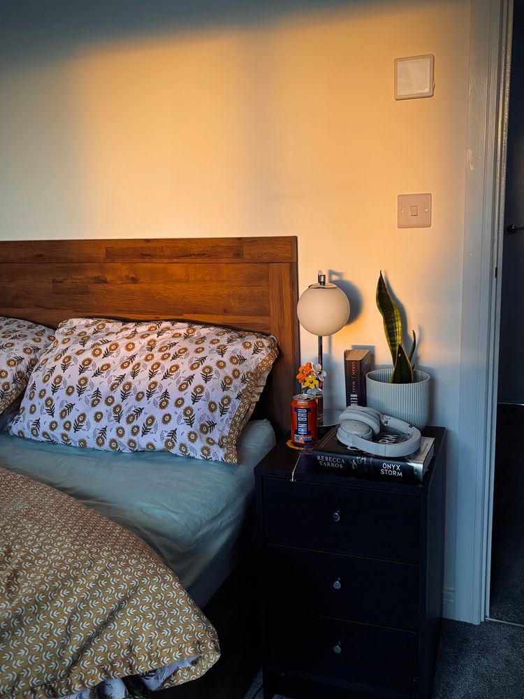 a low exposed image in golden hour, with the sunlight hitting the white wall of a bedroom. in the room is a wooden panel bed with a sunflower and leaf mustard duvet and green mattress covers. to the right is a black bedside unit with a white orb lamp, a marc jacob’s perfume, a can of irn bru on a crochet coaster, a Heir of Fire and Onyx Storm book, a snake plant in a white pot and a white bose headset.