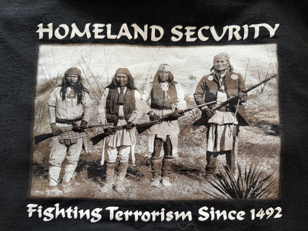 A black t-shirt with a photo of 4 indigenous men in traditional clothing carrying rifles. The shirt says “Homeland Security - Fighting Terrorism Since 1492”