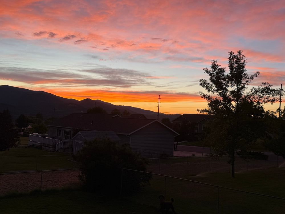 Pink fluffy clouds, a strip of blue sky and a bright orange cloud over a silhouetted mountain and house, and an aspen tree on the right. 
