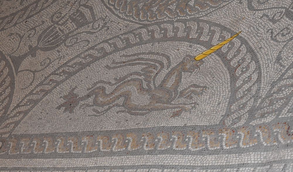 A Roman mosaic of a winged creature with a fish's tail and a felin head and fron paws, framed witin a half-circular twisted cord border. Some wag has placed a paper unicorn horn on the creature's head. 