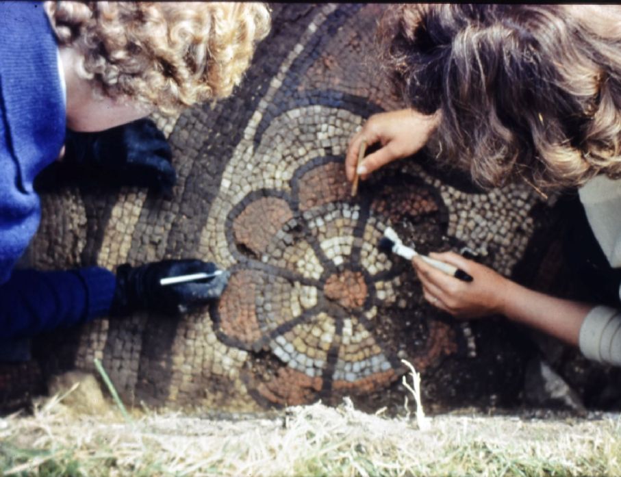 Overheat photo of the edge of an archaeological trench. At the bottom of the trench is part of a mosaic depicing a stylized flower. It is being carefully cleaned by two people. You can only see the backs of their heads and their hands. 