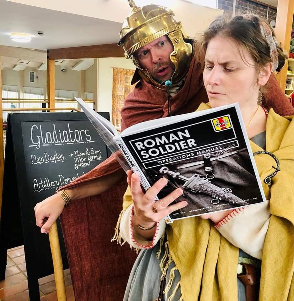 A celtic lady is reading a book (not an actual celtic lady, you understand, but someone dressed in celtic costume). The book is a Haynes manual-type of book called "Roman Soldier operations manual". Looking over her shoulder is a Roman soldier (or someone dressed as a Roman soldier, obviously). He looks slightly concerned. 