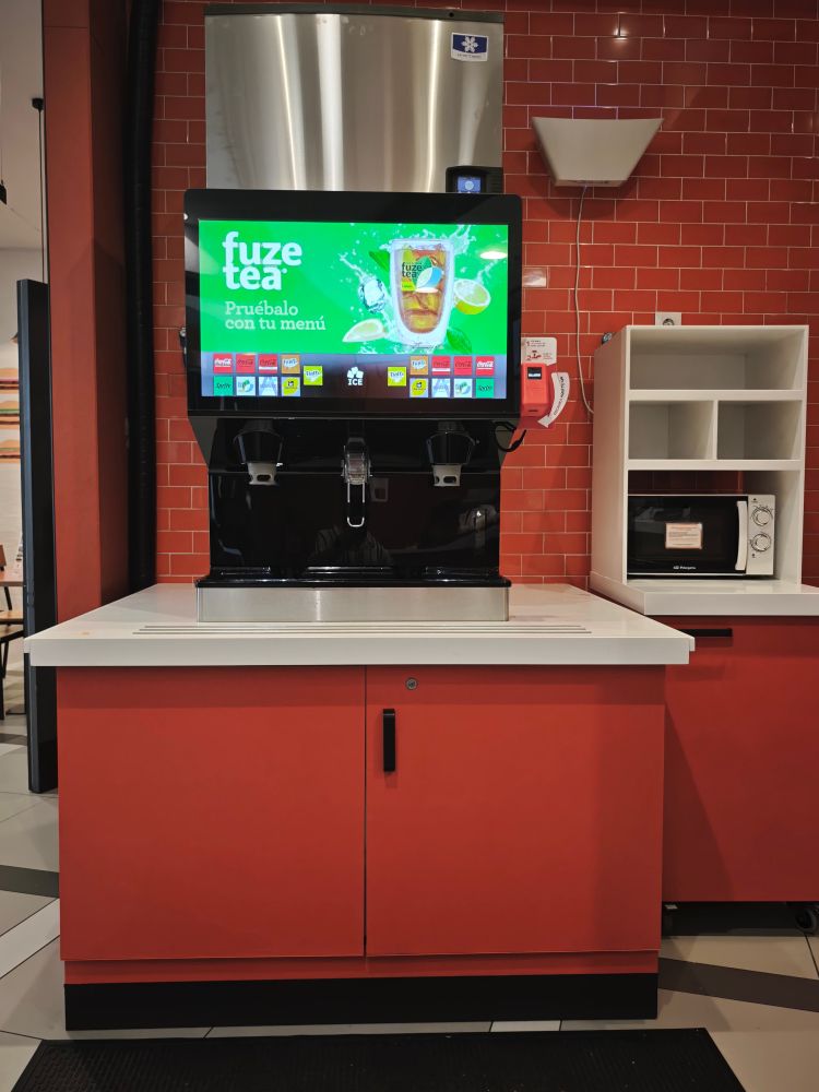 Nueva máquina de dispensación de bebidas del Burguer King.

Está encima de un mueble rojo con encimera blanca. La máquina no está pegada al borde por lo que hace que acceder a ella sea complicado 

Cuenta con una pantalla táctil sin ninguna herramienta de accesibilidad como un lector de pantalla.

Para poder usar la máquina es necesario usar un lector de códigos QR que está a la derecha y a una altura de 1,30 metros