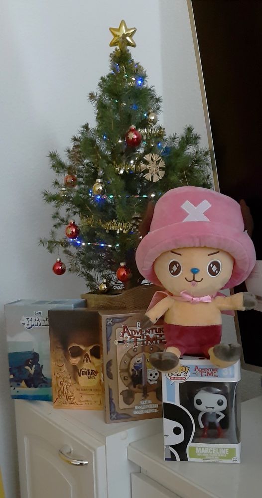 A photo of a small christmas tree. Underneath the tree are the complete DVD collections of Steven Universe, Venture Bros and Adventure Time. On the side there are also a Funko pop of Marceline from Adventure Time and a plushie of Chopper from One Piece.