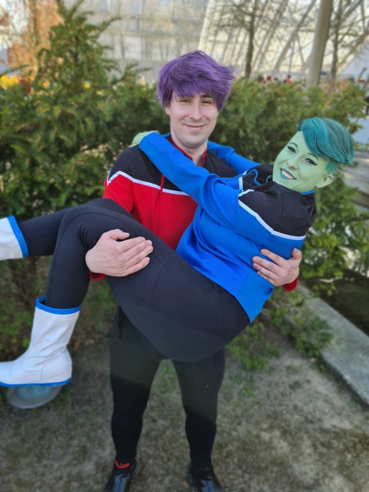 A photo of two cosplayers, portraying Bradward Boimler (red shirt) and D'Vana Tendi (blue shirt) from Star Trek: Lower Decks. Boimler is lifting Tendi bridal style, they look happy.