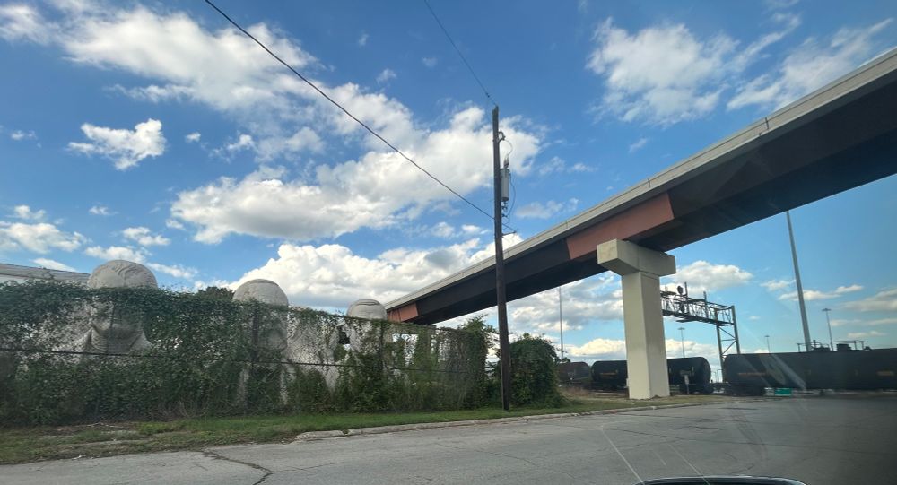 view from a stopped car: a line of tank cars under an overpass next to David Adickes’s large white sculptures of president heads behind a vine-covered fence