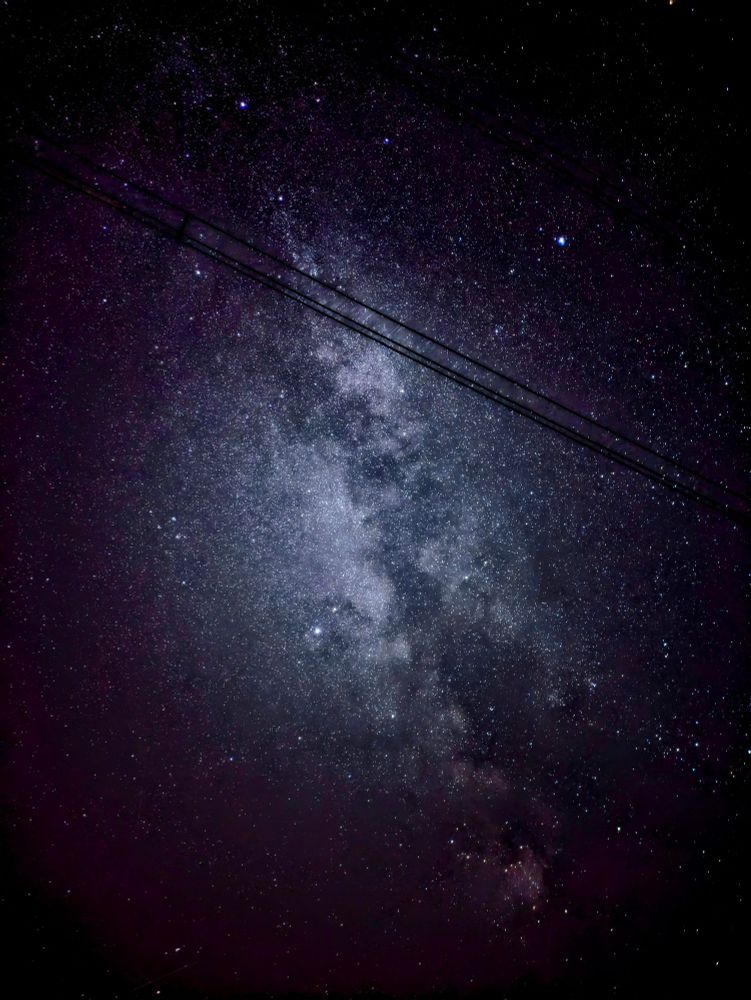 Camera: Google Pixel 7a
Adjustmemts made by me. 

The image depicts a night sky filled with countless stars. The most prominent feature is a band of light stretching across the sky, which is likely the Milky Way galaxy. This cosmic band is characterized by its diffuse, milky appearance and its concentration of stars compared to the surrounding areas. The night sky is mostly dark, with a few brighter stars visible.
In the foreground, there is a power line cutting through the image vertically, creating a contrast between the natural beauty of the night sky and the man-made structure. The power line serves as a reminder of human presence and technology amidst the vastness of the cosmos.