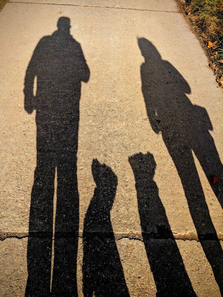 In diesem Bild sind vier verlängerte Schattensilhouetten nebeneinander auf einem Steinboden zu sehen. Die beiden äußeren sind die von der Frau (rechts) und mir (links). Zwischen uns sind die Schatten der Pudelmädels, welche jeweils ihren Blick in die Richtung von einem von uns haben. 