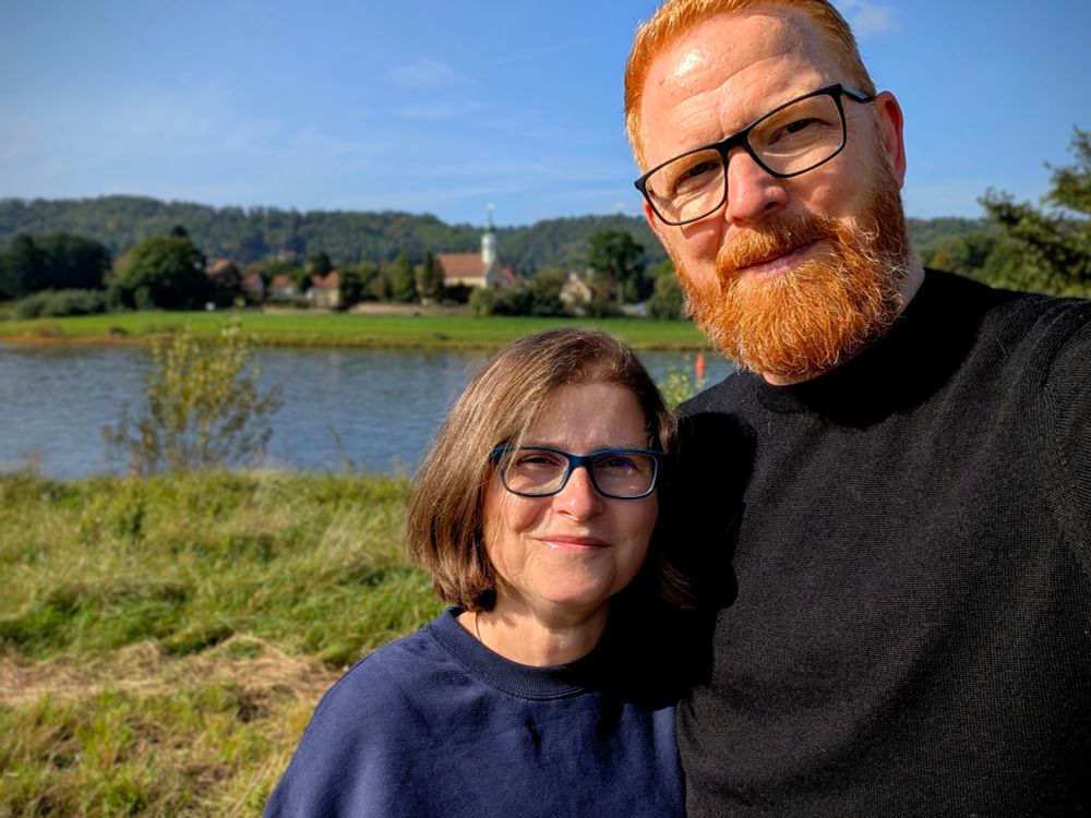 Um rechten Bereich des Bildes eine  Frontalaufnahme von mir und meiner Frau, links davon, leicht verschwommen ein Stück der Elbwiese, die Elbe selbst und am anderen Ufer ehe kleine gelb-weiße Kirche mit roten Ziegeln. 