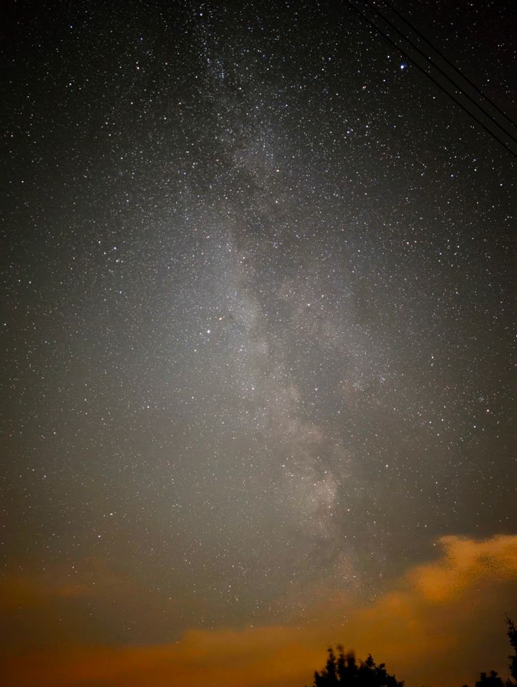Das Bild zeigt eine beeindruckende Langzeitbelichtung des Nachthimmels. Im Zentrum des Bildes erstreckt sich die milchig-weiße Struktur der Milchstraße. Sie verläuft von oben nach unten durch die Mitte des Bildes und ist umgeben von unzähligen hellen und schwächeren Sternen, die über den gesamten Himmel verteilt sind.
Am unteren Rand des Bildes sind diffuse, orangefarbene Wolken zu sehen, die vermutlich durch die Lichtverschmutzung einer Stadt oder einer anderen künstlichen Lichtquelle von unten beleuchtet werden. Unter diesen Wolken sind die dunklen Umrisse von Bäumen oder Sträuchern erkennbar. Am oberen rechten Rand des Bildes sind zudem einige dünne, dunkle Linien zu sehen, die Stromleitungen sein könnten. Die Aufnahme wurde wahrscheinlich außerhalb des Stadtzentrums gemacht, um einen so klaren Blick auf die Milchstraße zu erhalten, da die Lichtverschmutzung innerhalb von Städten dies normalerweise verhindert.
