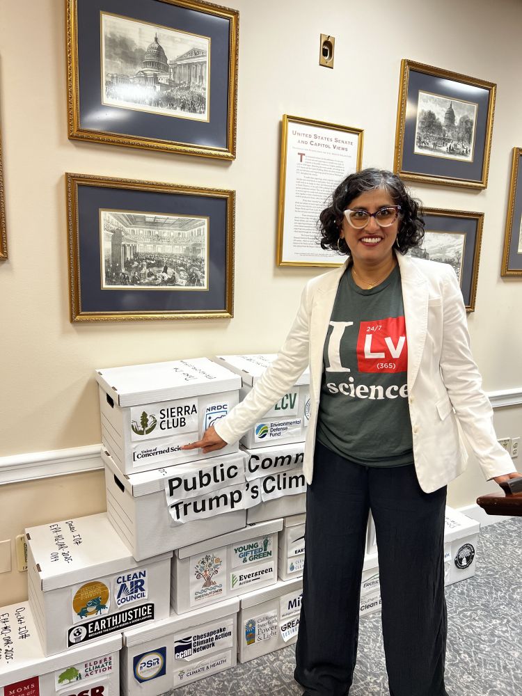 A woman wearing a white lab coat and glasses stands near several file boxes marked "Public Comment" with various environmental organizations' logos on them. 