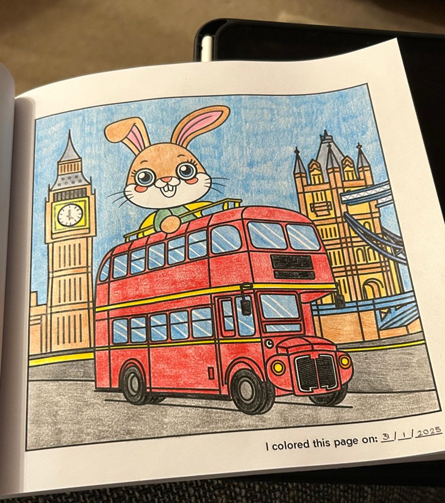 A colouring in of a London bus with a cartoon rabbit sitting on the top deck in the open air. The bus is passing the Houses of Parliament and Tower Bridge. It is an imagined version of London. 