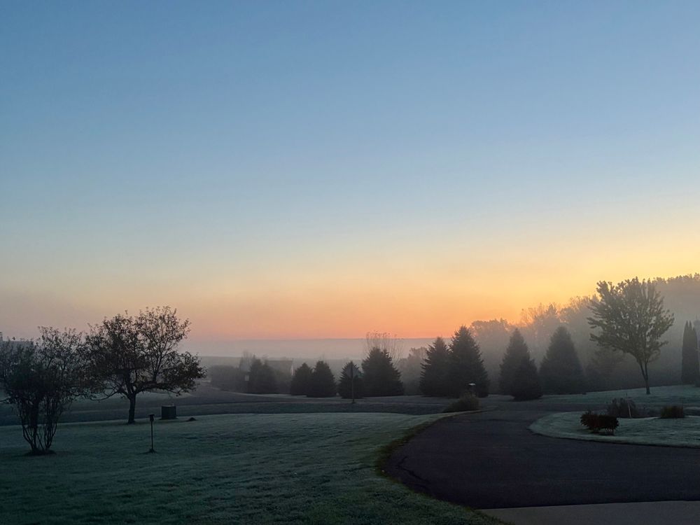 Misty orange and blue sunrise sky, with evergreens and apple trees  in the near distance. 
