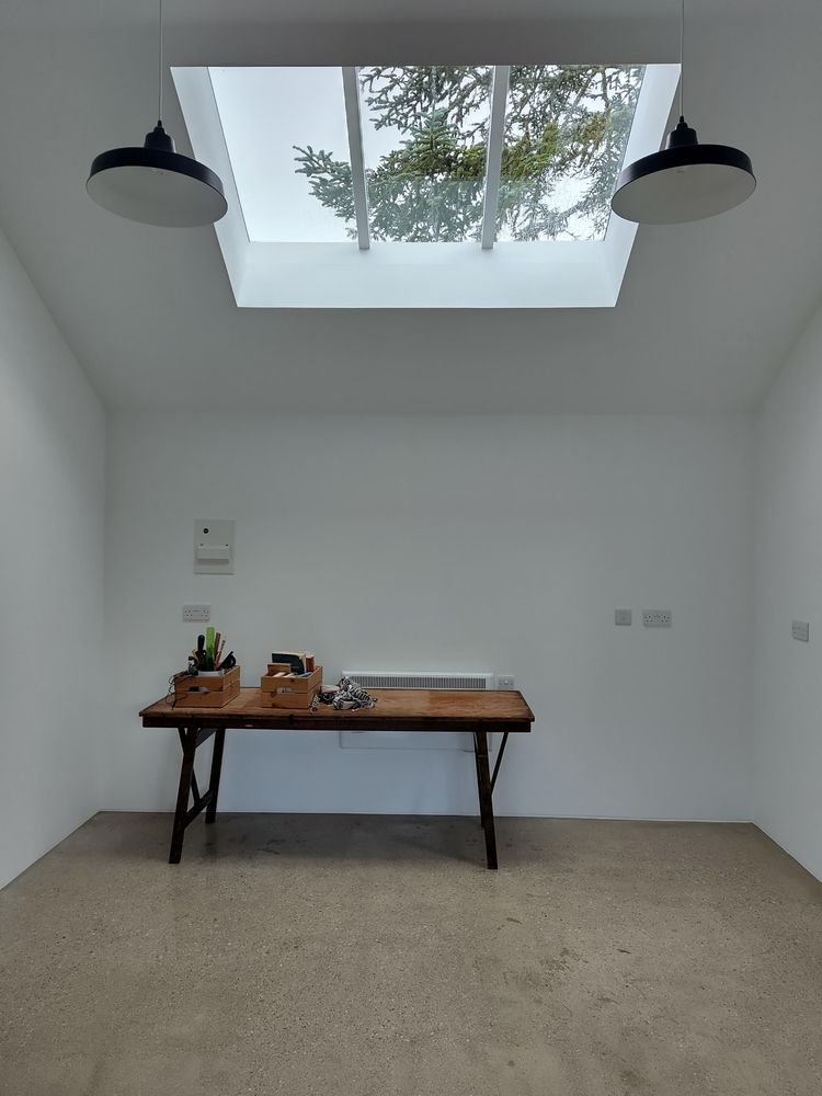 A long wood table in an otherwise empty room. The space has a sloped ceiling with a large roof window and a tree visible above. There are two simple black and white pendant lights. It is painted white, with a polished concrete floor.
