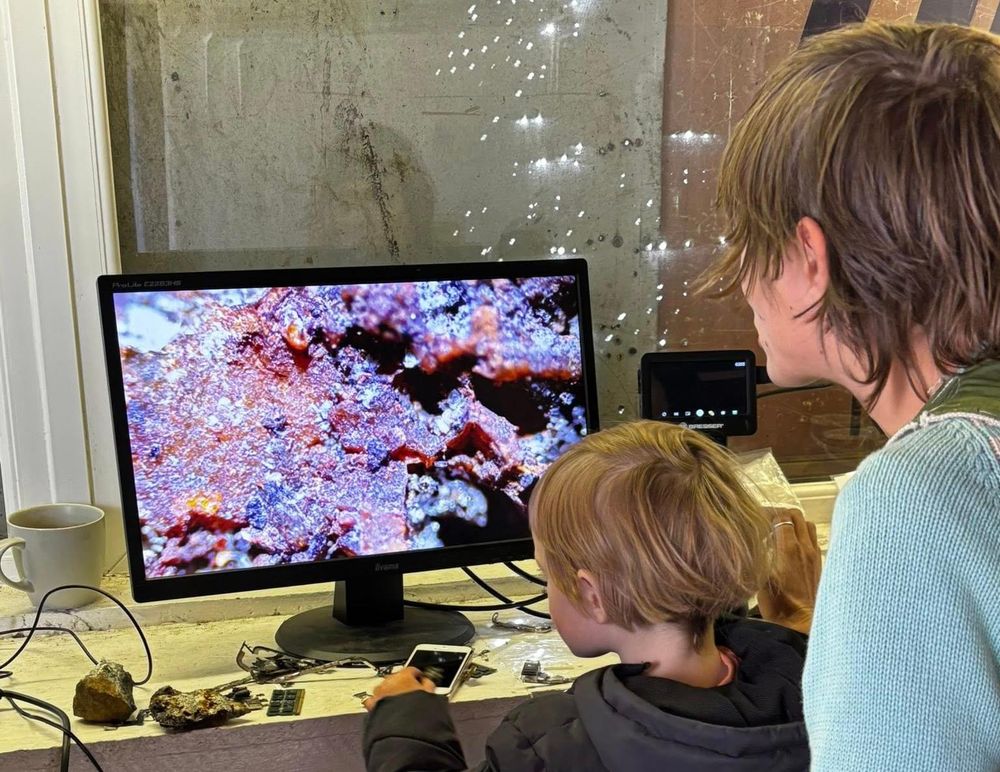 Two people looking at a microscope image on a monitor with scattered rocks and e-waste on the table.