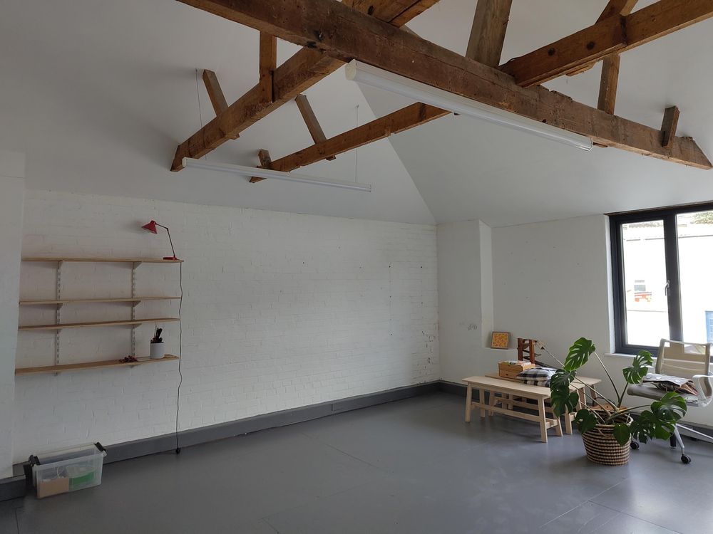 Almost empty new studio with grey painted floor, a few shelves on the wall, and big wooden beams with a high ceiling. A few bits have been moved in including a monstera plant and some wood benches.