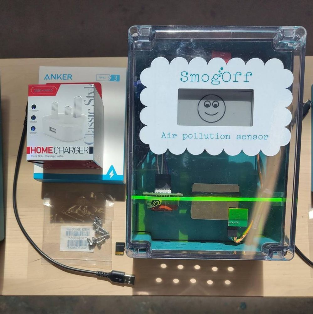 A smogoff sensor viewed from the front, with a smiley face showing on the screen, a cloud-ish shaped sticker around the screen, and a fluorescent acrylic shelf inside holding the various sensors