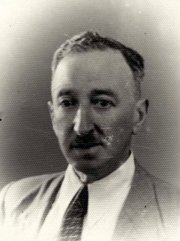 Black and white portrait of a man with moustache wearing a suit and tie, looking directly at the camera with a neutral expression.