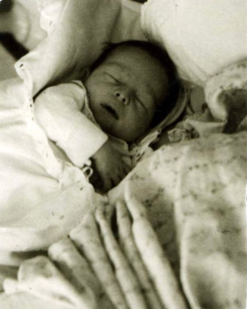 A baby girl photographed from above. She is sleeping. She is covered with a blanket and surrounded with pillows. Her mouth is open.