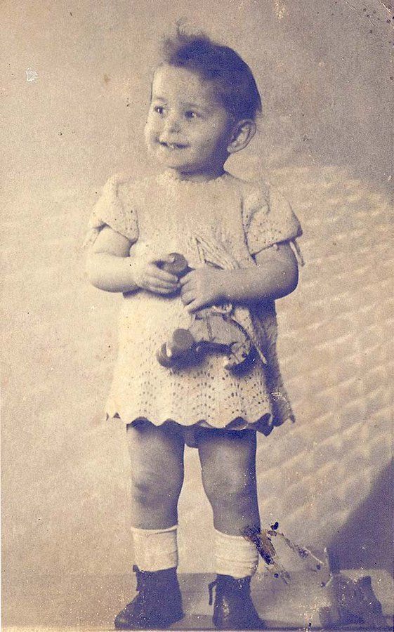 Vintage black-and-white photo of a young girl standing and holding a stuffed toy. The child is wearing a short-sleeved, knee-length dress with a textured pattern, socks, and shoes.