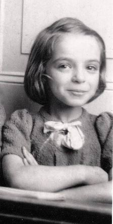 A vintage black and white photo of a young girl smiling, wearing a sweater with a bow on the collar, arms crossed in front of her.