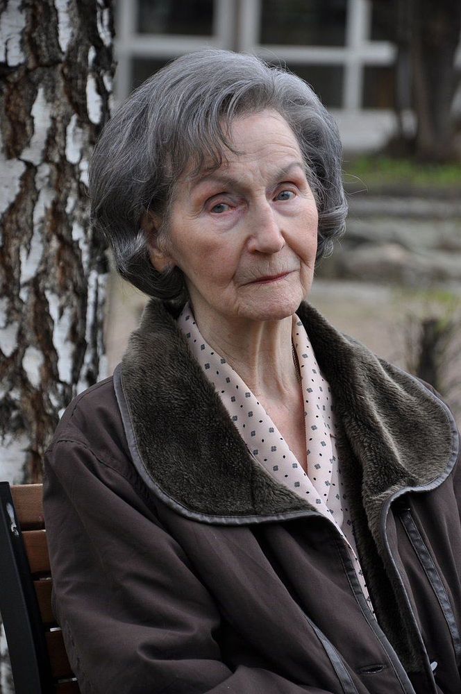 A senior woman with gray hair sits on a bench outdoors, leaning against a tree, wearing a jacket with a fur collar and a polka-dot blouse, displaying a thoughtful expression.