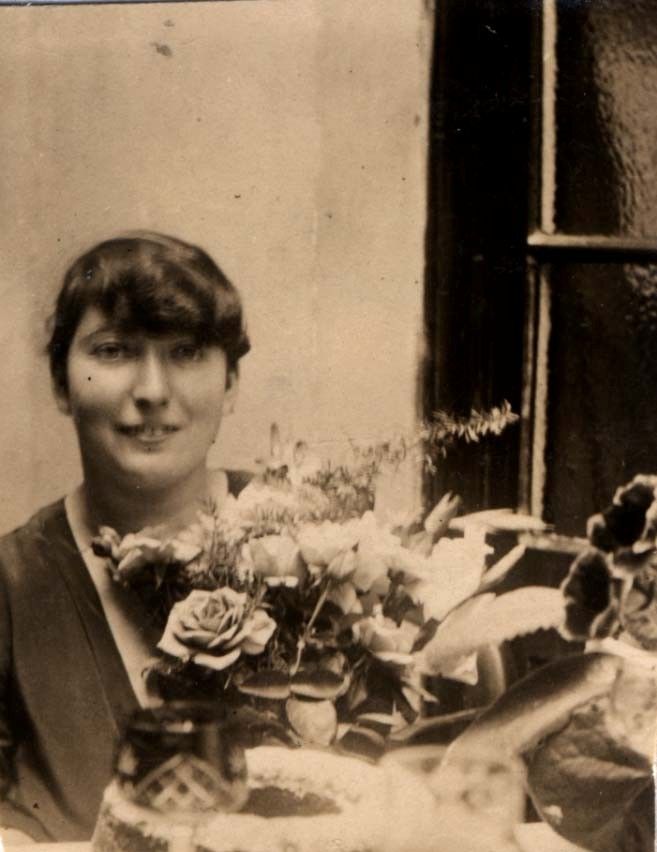 Sepia-toned vintage photograph of a smiling woman seated next to a window, surrounded by an array of flowers.