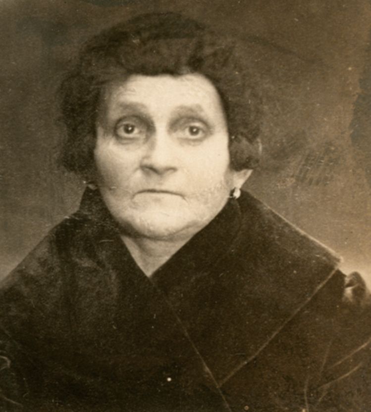 Vintage black-and-white portrait of an elderly woman with a solemn expression, wearing a dark, high-collared coat and styled hair. The photograph has a faded and time-worn appearance.