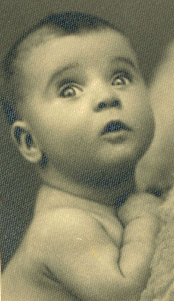 Close-up photo of a baby with wide-open eyes and a slightly open mouth, looking upward with an expression of surprise or curiosity. The image is in sepia tone, suggesting an older photograph.