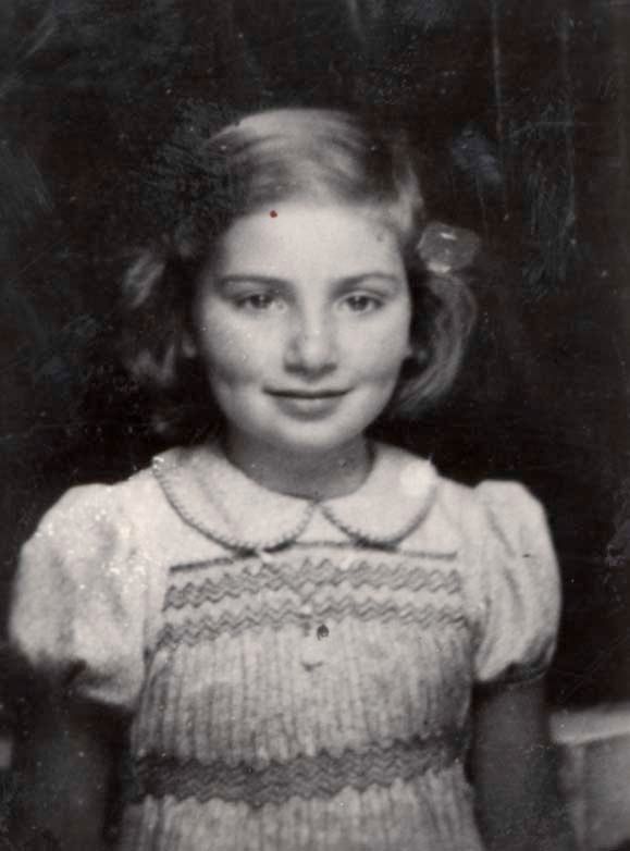 Portrait photograph of a young girl. She is wearing a short-sleeved dress with a collar with horizontal patterns. Around her neck is a pendant. Her long hair is styled in a ponytail. She is smiling slightly.
