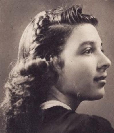 A photo of the face and shoulders of a young woman. She is photographed from the back and she turns her head to show her profile. She has ling hair styled waves and curls.