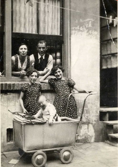 Two adults and three children posing in front of a window, one of the children is sitting in a vintage pram, against a backdrop of a weathered building facade