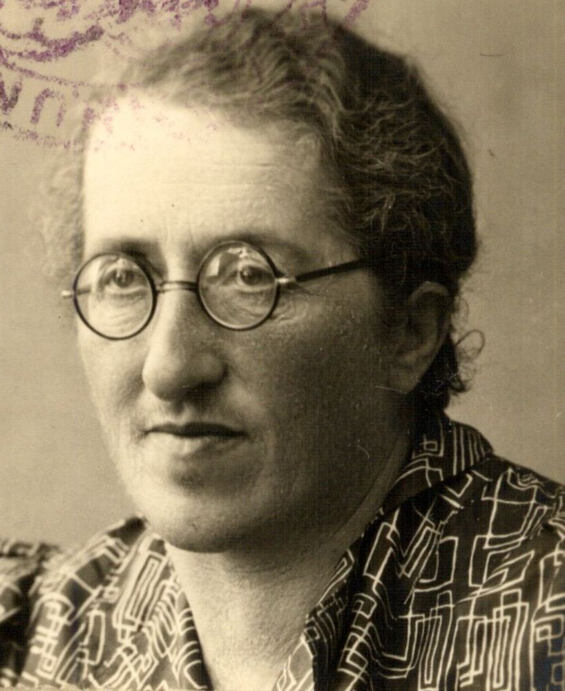 Vintage sepia-toned portrait of Emma Goldman, featuring her wearing round glasses and a patterned blouse, looking slightly off-camera with a thoughtful expression.