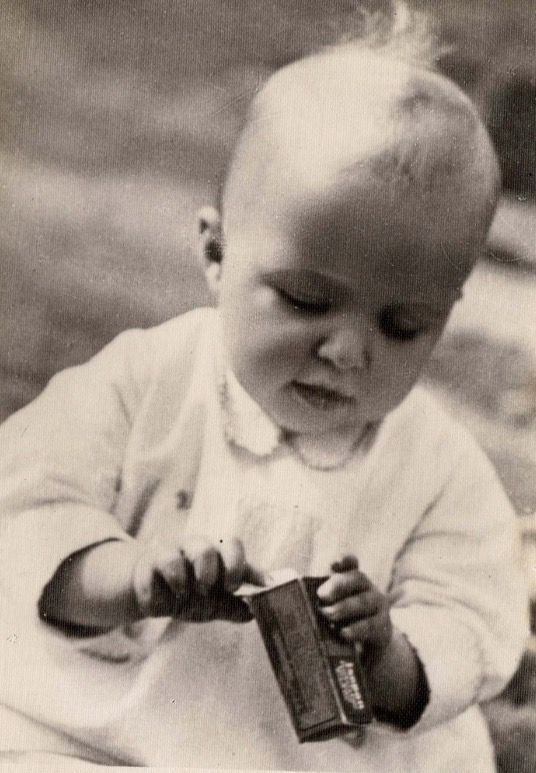 A baby girl photographed outside. She has a bit of blond hair at the top of her head. She is wearing bright clothes and is busy holding some box in her hands.