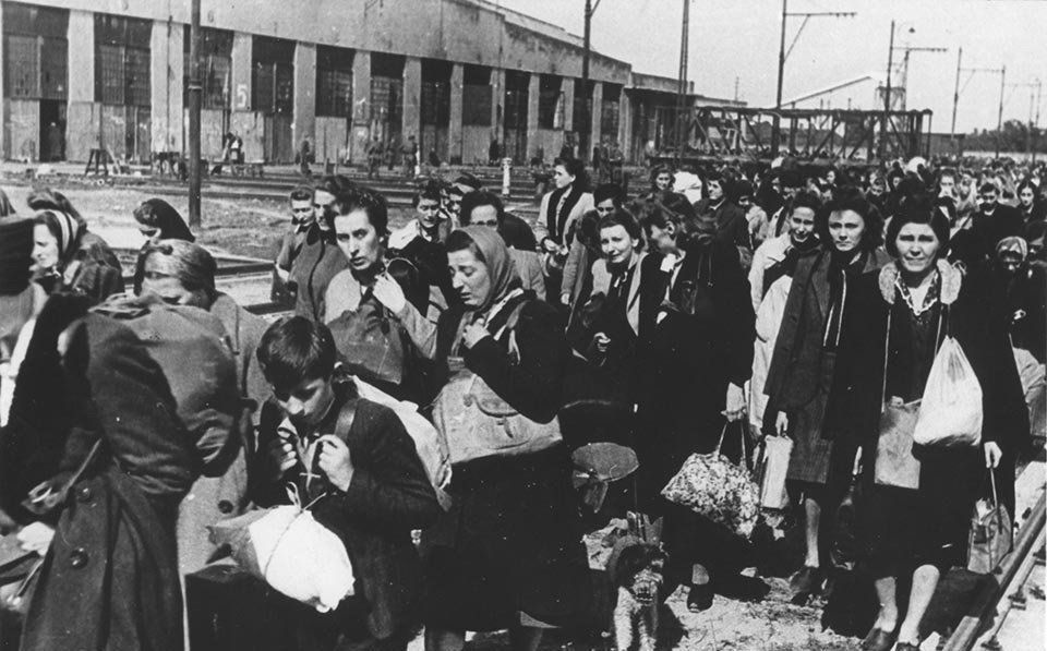 Group of people - mainly women and children - walking along a rail line. A large building in the background. Inhabitants of Warsaw deported from the Pruszków camp during the Warsaw Uprising.