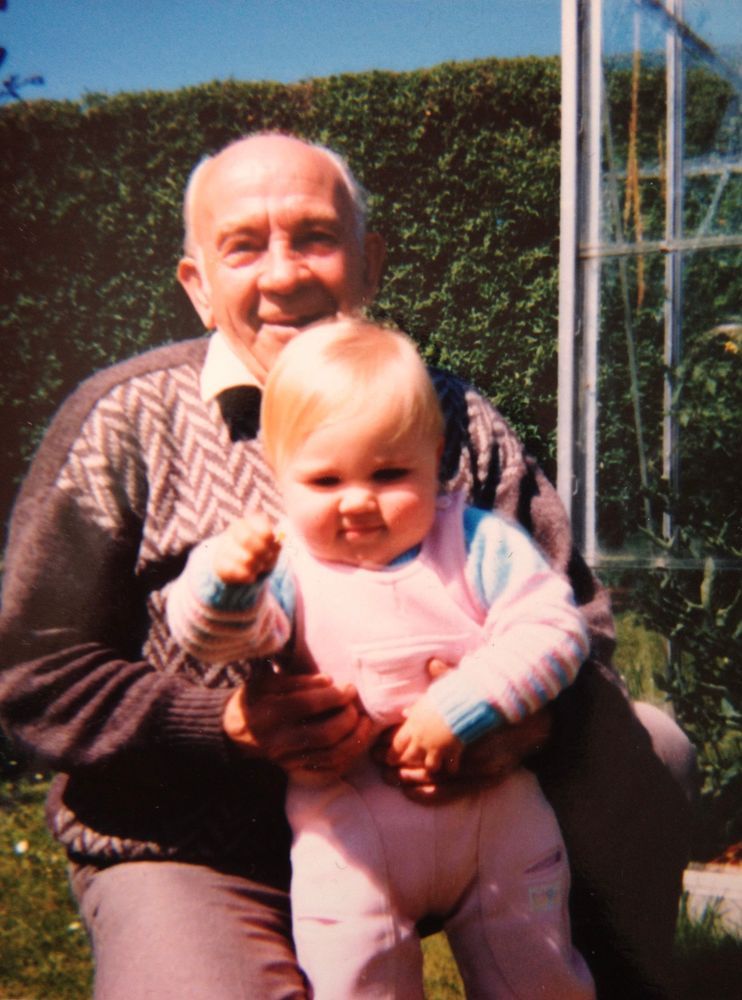 A color photograph of an elderly man holding a young baby, both smiling warmly. The man has gray hair and is wearing a patterned sweater, while the baby is dressed in a light pink outfit with blue sleeves. They are sitting outside in a garden, with a lush green hedge and a glass structure visible in the background.