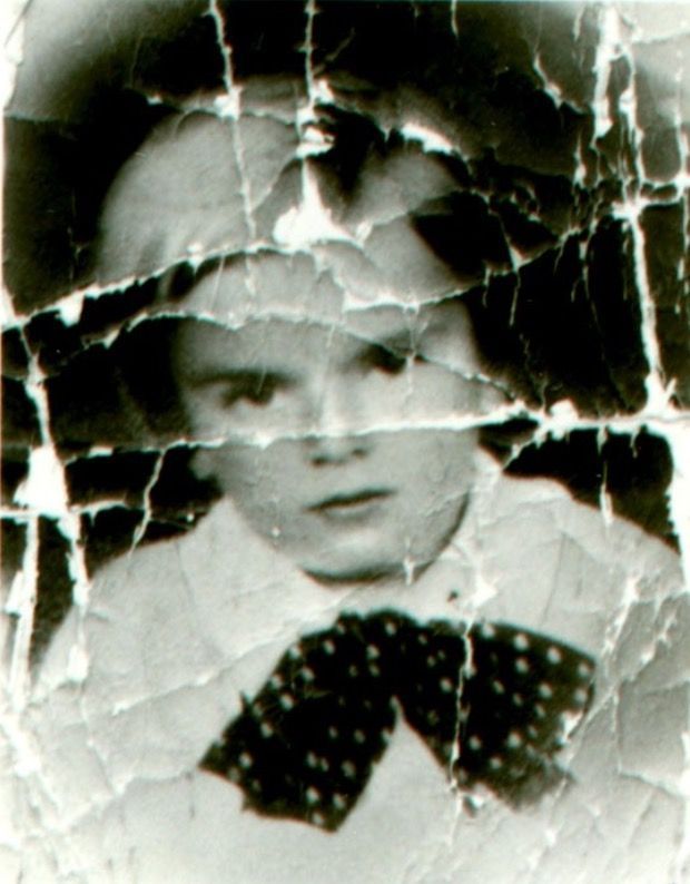 Picture of a young boy's face. He is wearing a shirt and a big polka dot bow under his neck. He has slightly longer hair pulled back to the right side. The photograph is tattered - you can see the marks and losses from the bends.