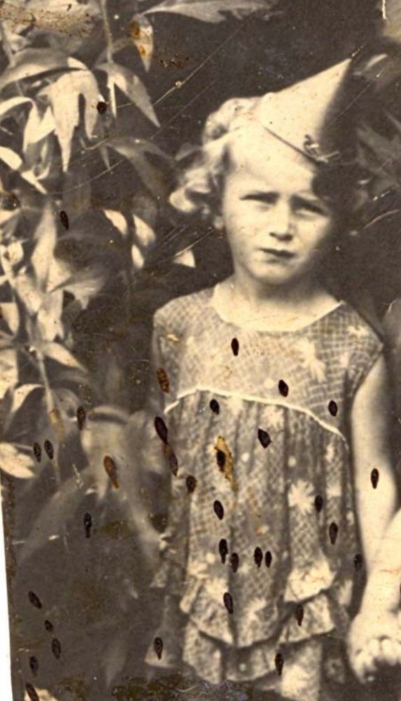 Vintage photograph of a girl wearing a party hat and a patterned outfit, standing in front of leafy foliage.