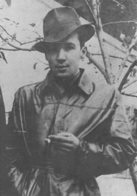 Black and white photograph of a person wearing a fedora and a trench coat, holding a cigarette in one hand, standing beside a leafless tree.