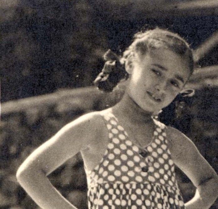 Vintage black and white photo of a young gril in a polka-dot dress, standing with hands on hips and looking slightly to the side.