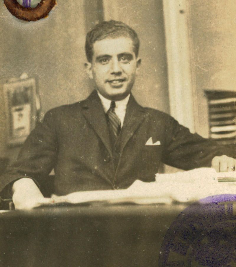 Vintage sepia-toned photograph of a man sitting at a desk with papers, looking directly at the camera.