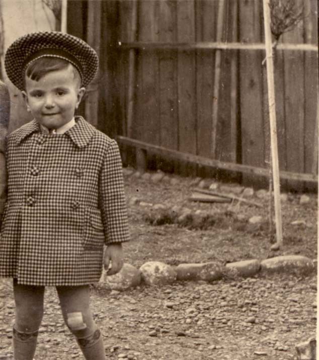 A boy in a checkered coat and hat standing outdoors with a faint smile, with a wooden fence and bare trees in the background.