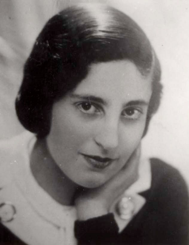 A close-up portrait of a woman with a focused expression. She wears a light-colored blouse with a dark lapel and a floral pin. She rests her head onto her hand.