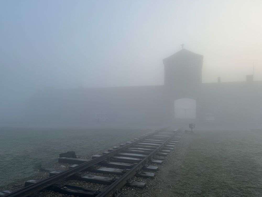 A foggy morning view of the railway tracks leading to the entrance gate of Auschwitz II-Birkenau. The iconic brick gatehouse is barely visible through the thick mist, creating an atmosphere of somber stillness and reflection.